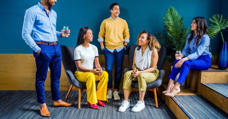 A diverse group of five professionals engaging in a casual indoor meeting, smiling and talking.
