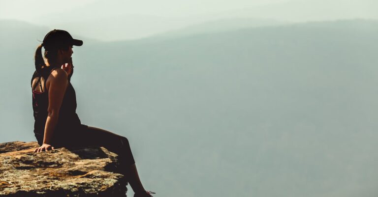 A woman enjoys a calm moment sitting on a cliff's edge against a foggy mountainous backdrop.