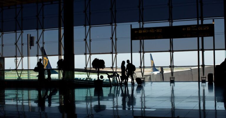 Travelers silhouetted in a modern airport terminal with glass windows and visible airplane tails.