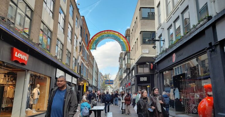 A bustling urban street with shops and a colorful rainbow decoration under a bright blue sky.