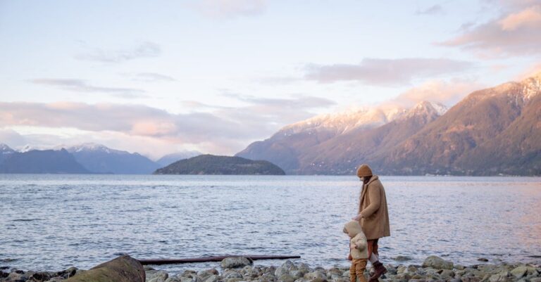 Side view of mother and little kid in casual warm outwear walking together along calm water on stony shore and holding hands