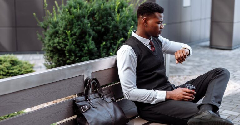 Young professional sitting on a bench, checking his wristwatch with coffee in hand.