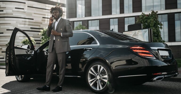 Elegantly dressed man in gray suit making phone call next to sleek black car.