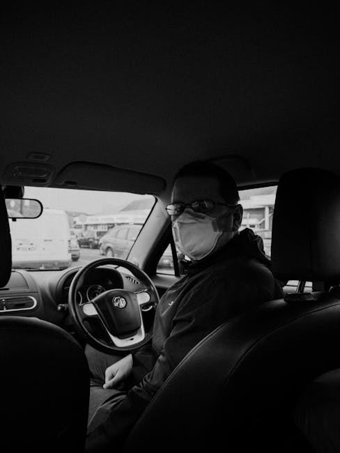 Man sitting inside a vehicle wearing a face mask, in grayscale.